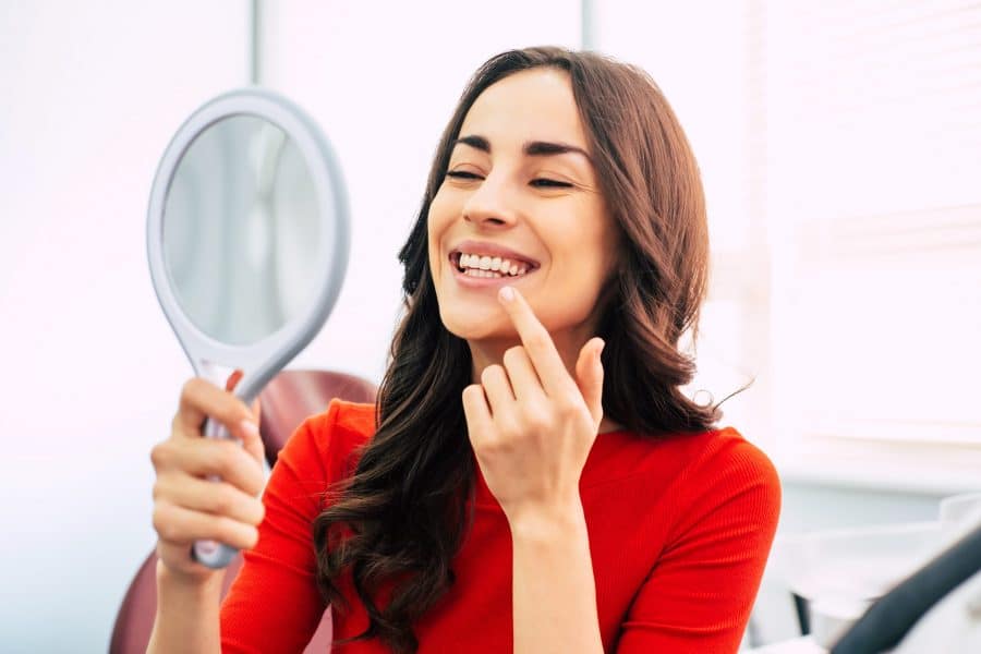 Brunette woman in a red shirt smiles and looks into a handheld mirror