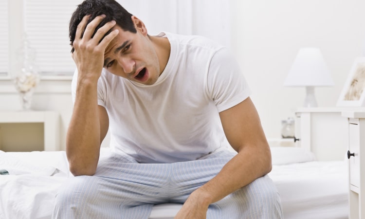 Brunette man in his pajamas yawns and holds his forehead because he has a headache and is tired due to obstructive sleep apnea