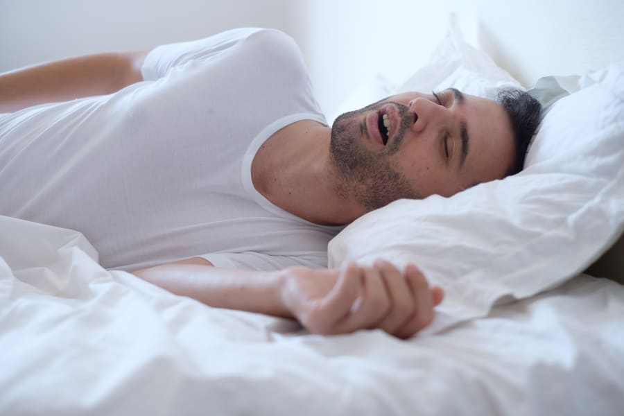 A brunette man snores as he sleeps on his back in his bed