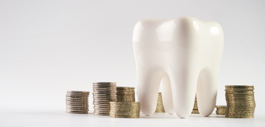 A large model of a white tooth next to stacks of coins against a gray background